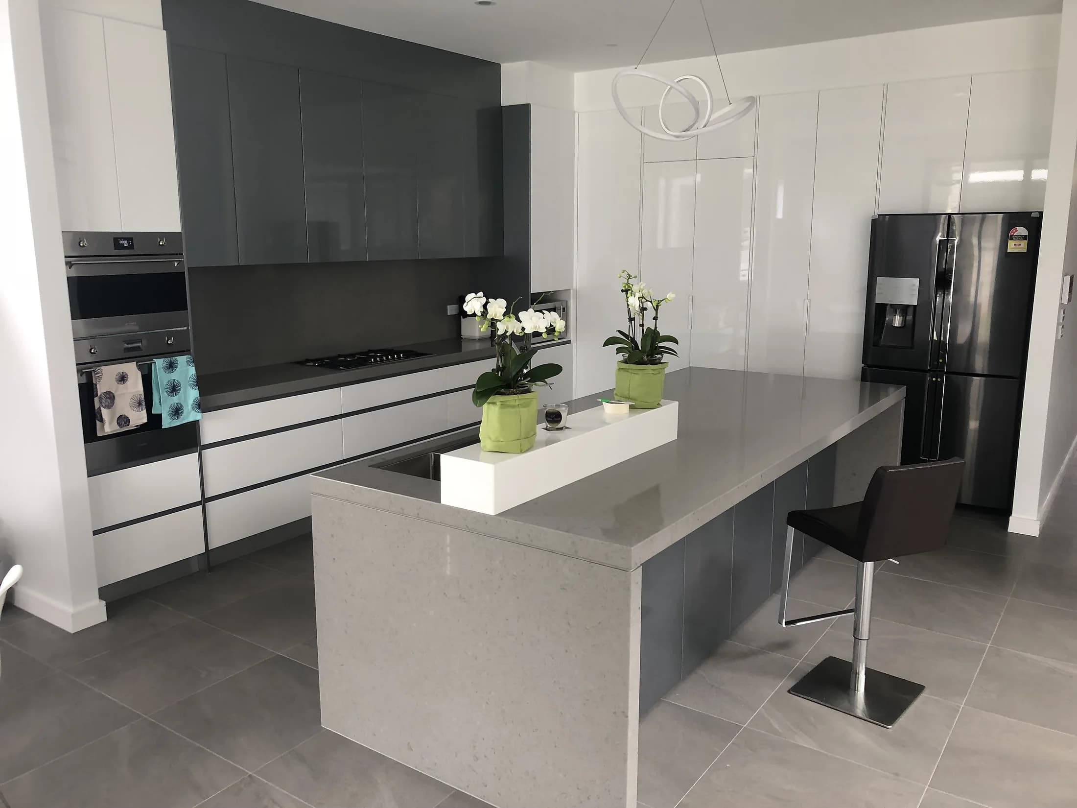 White and charcoal handleless kitchen with a grey stone-look island, wall ovens and a tall pantry wall, by H&R Kitchens