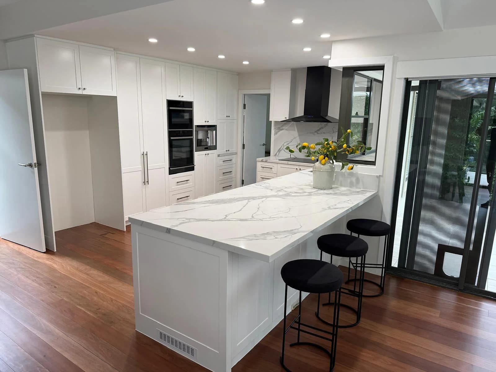 Bright white shaker kitchen with a large marble-look island, black rangehood and black bar stools, by H&R Kitchens