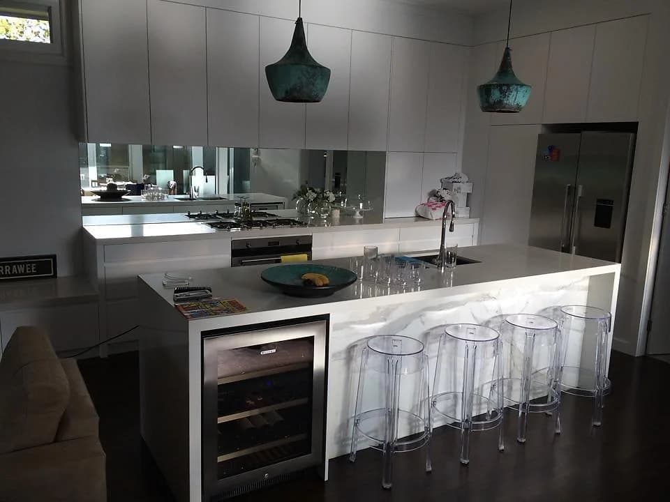 Open-plan white kitchen with a mirrored splashback, a waterfall island with a wine fridge and teal pendant lights, designed by H&R Kitchens
